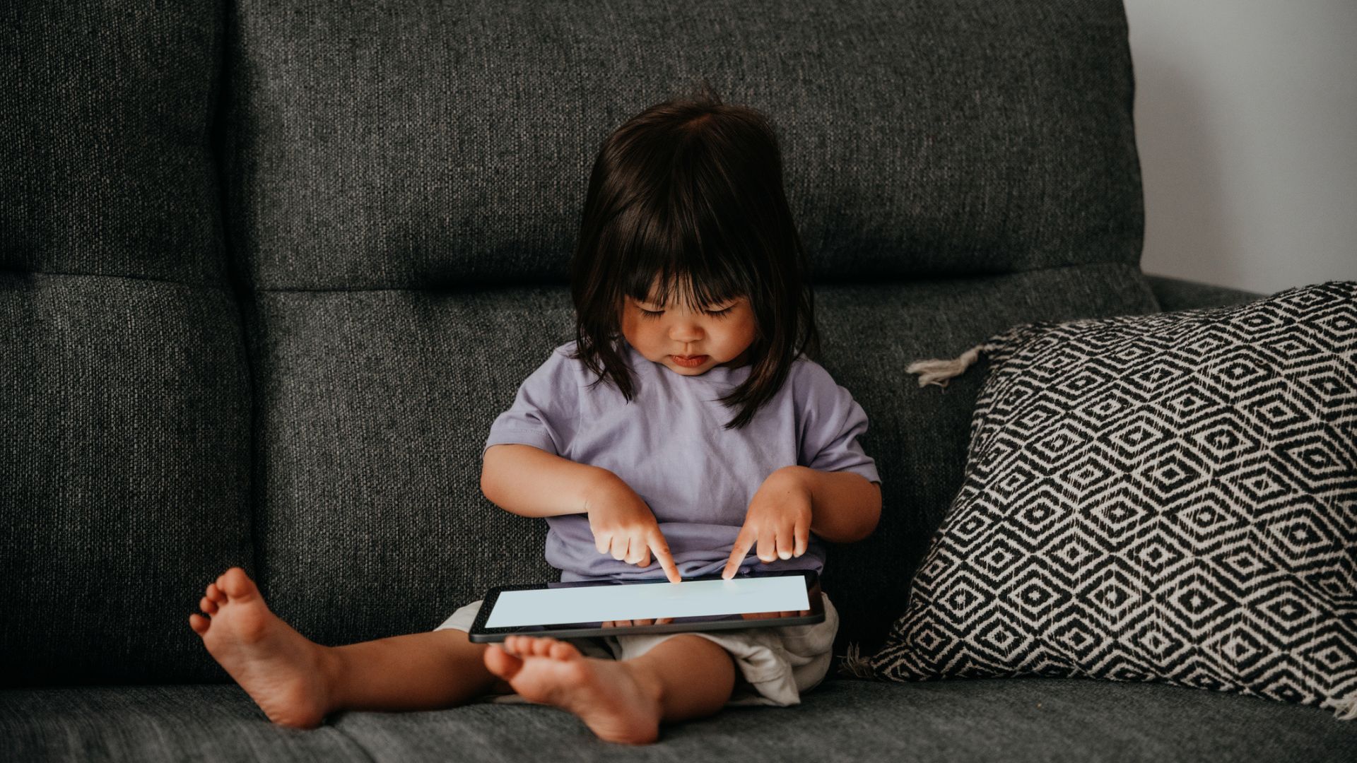 A toddler sits on a sofa with an iPad on her lap