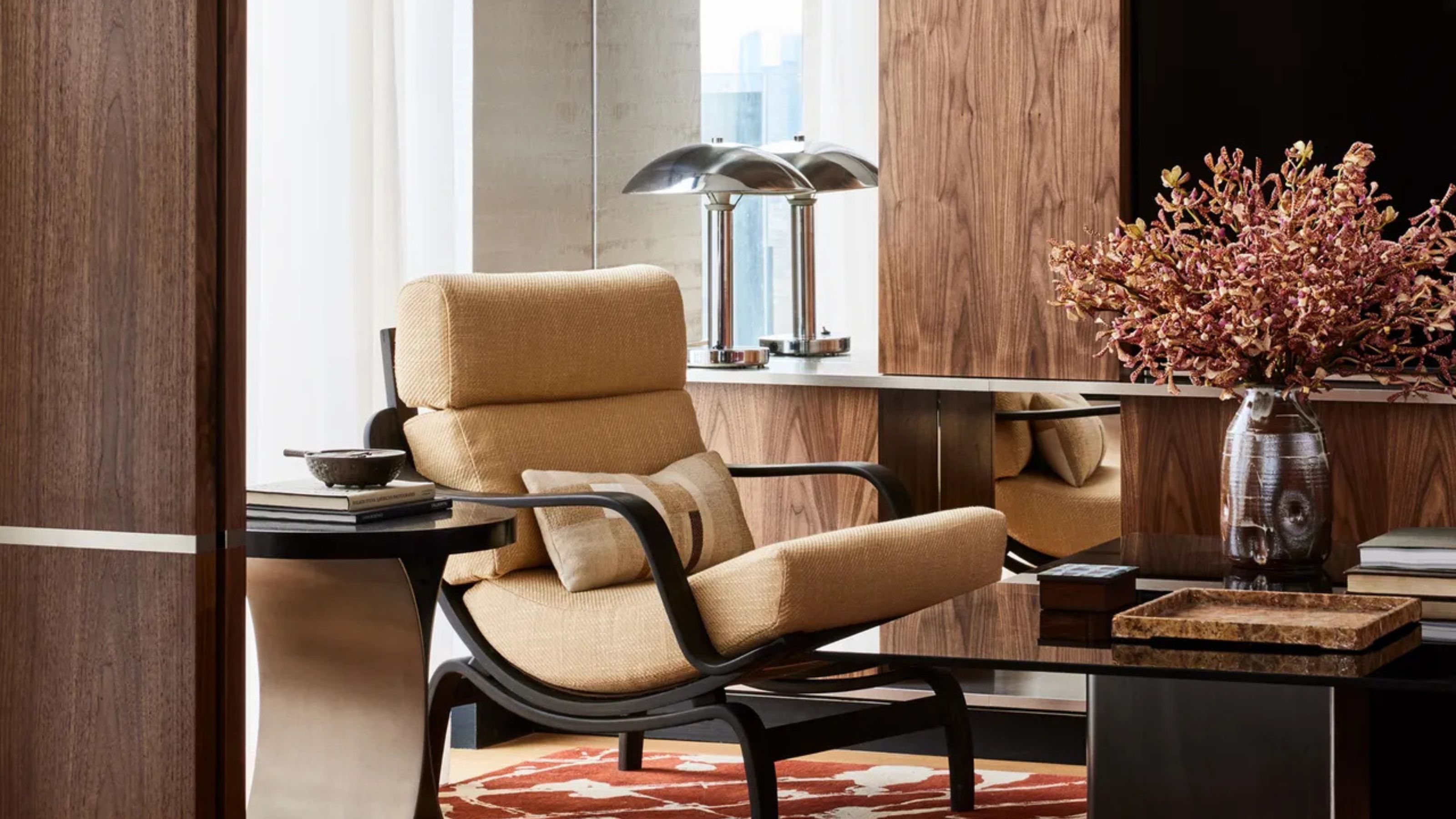 living room with armchair, coffee table styled with books and a vase of flowers, side table next to the chair, and then a built-in unit with mirrored backsplash reflecting a chrome lamp