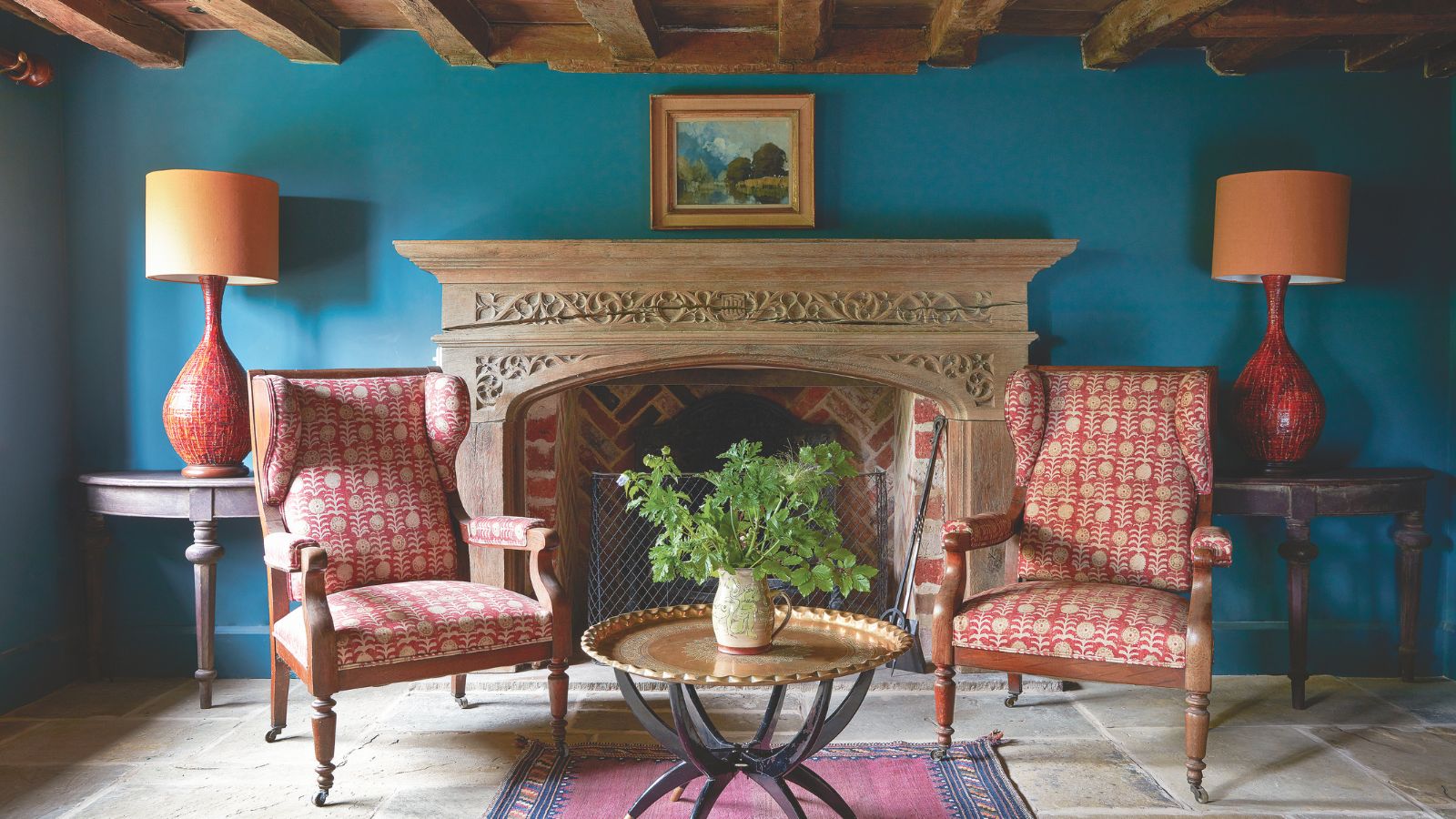 Colorful sitting room with two red patterned armchairs, an ornate fireplace, a side table with potted plant, a red rug, pictured in front of bright blue wall with wooden-beamed ceiling above.