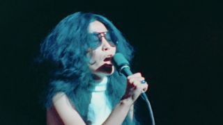 Yoko Ono wearing sunglasses and singing into a microphone
