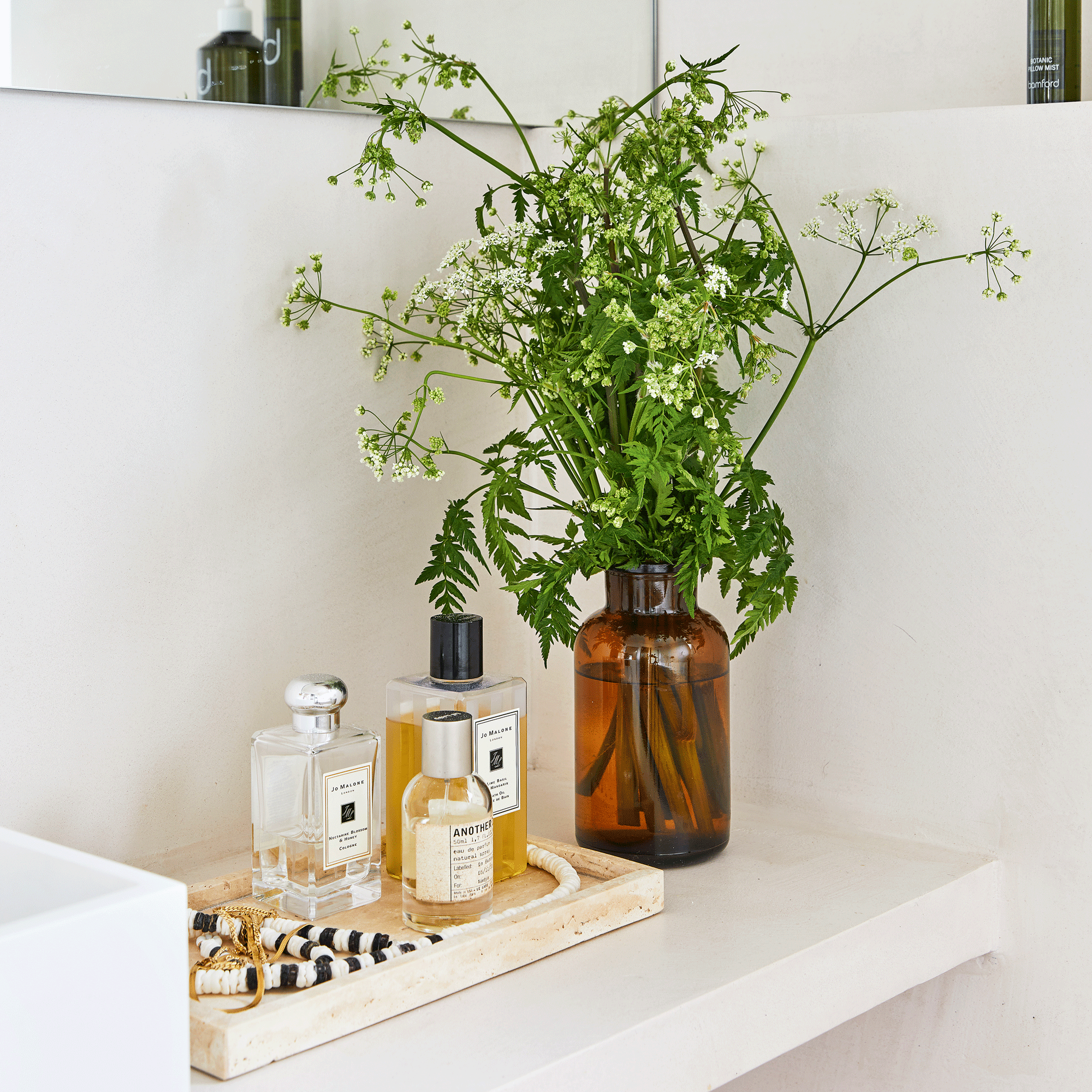 a bathroom countertop with a trinket tray filled with Jo Malone and Le Labo perfume and toiletries beside a bottle vase filled with flowers and greenery