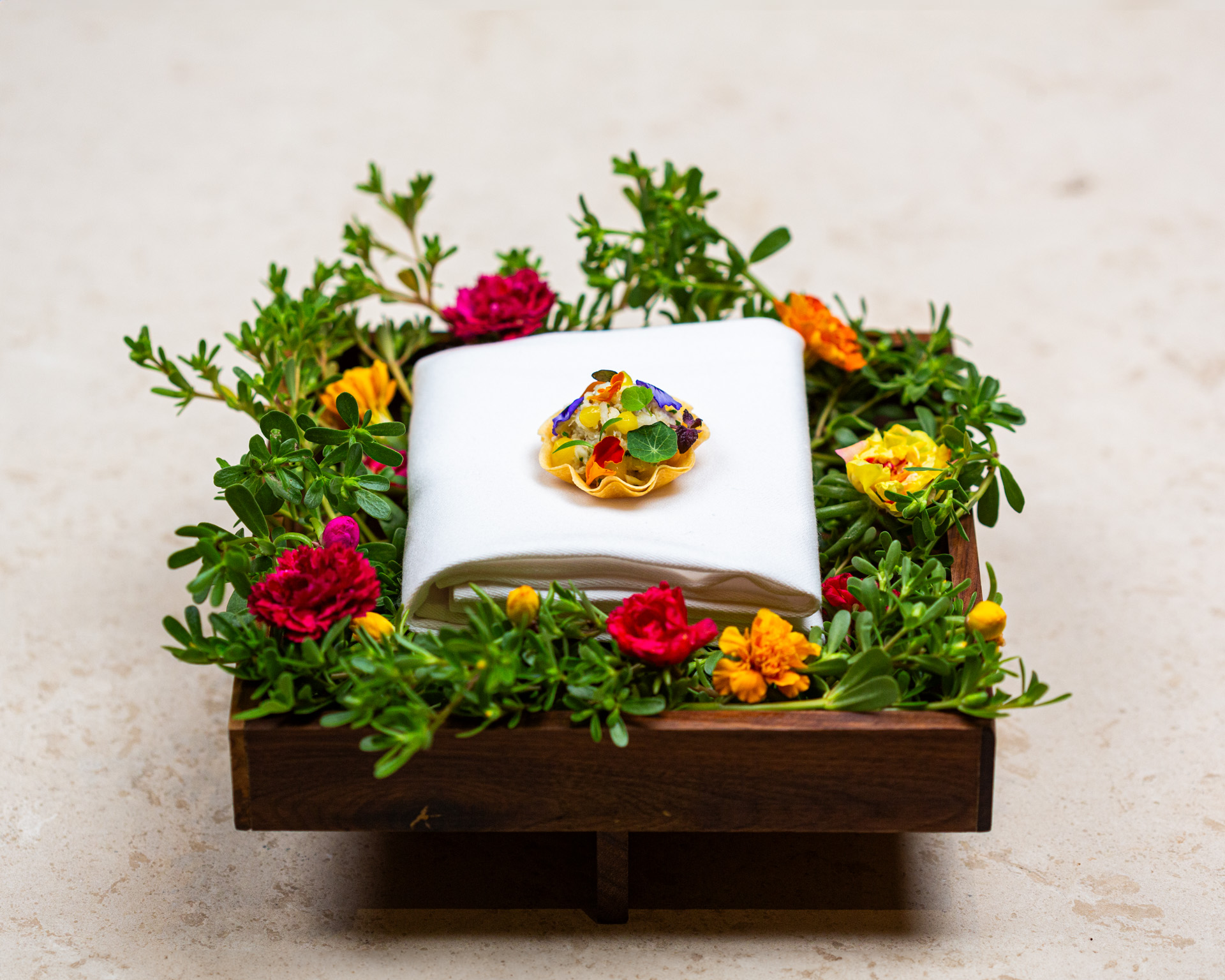 A crab tartlet beautifully plated atop a white cloth napkin surrounded by flowers and greenery in a wooden tray set atop a stone surface.