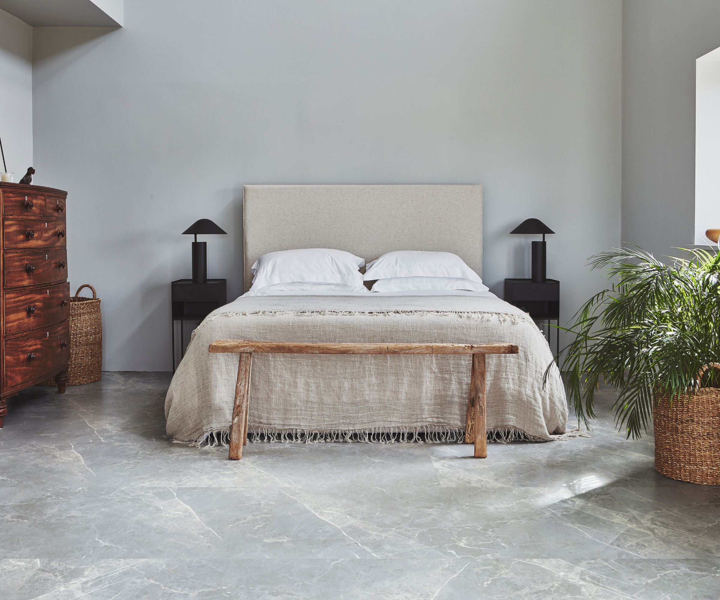 A calm, minimal bedroom with large-format grey marble-effect floor tiles and soft grey walls. A linen upholstered headboard with white bedding and a fringed throw sits between two black bedside tables with matching lamps, with a rustic driftwood bench at the foot and a large potted palm alongside an antique mahogany chest of drawers.
