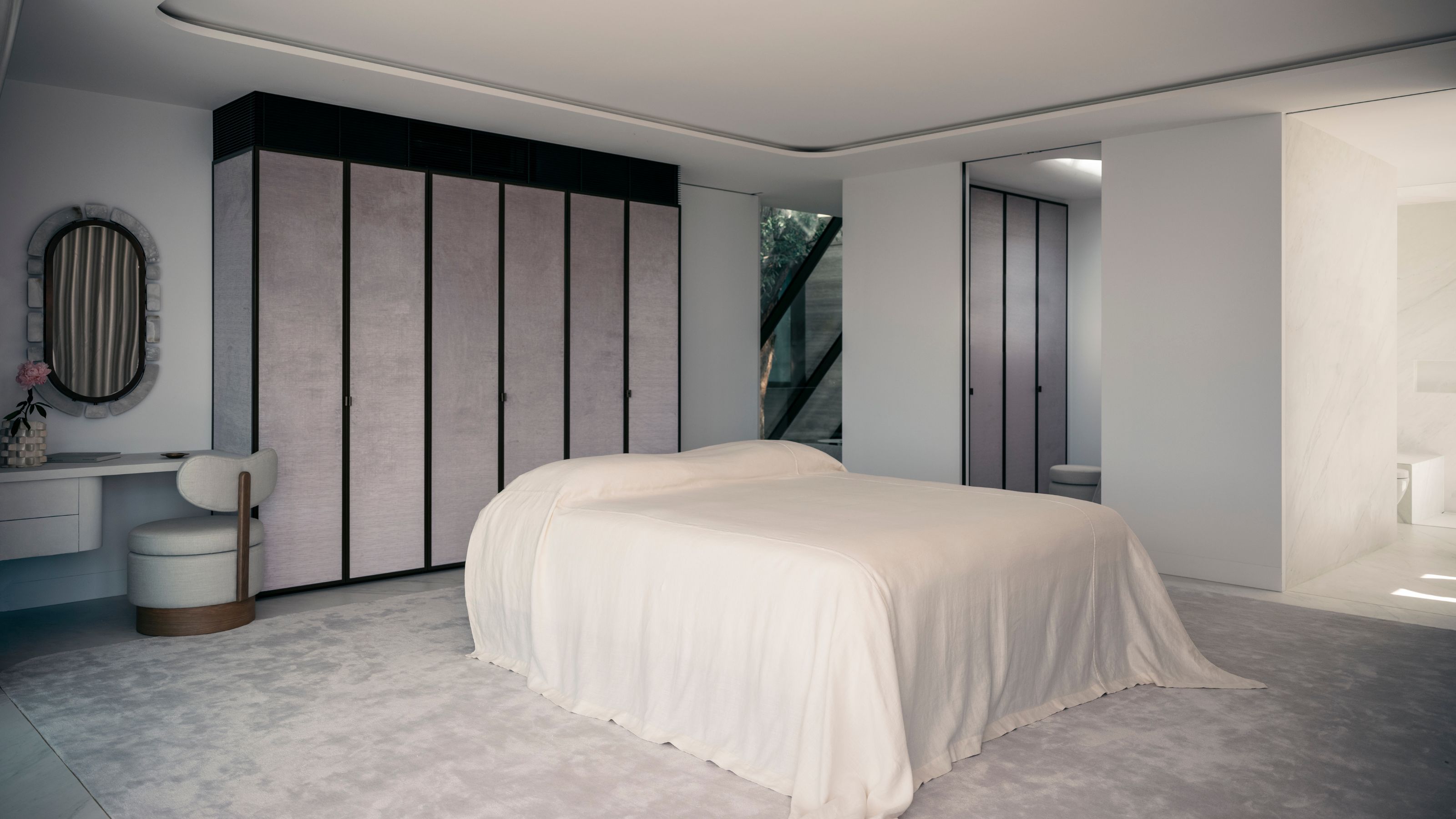 Image of a minimalist, luxury bedroom with gray-toned walls and a gray-toned carpet. There is a modern, minimalist bed with a white bedspread in the center of the room.