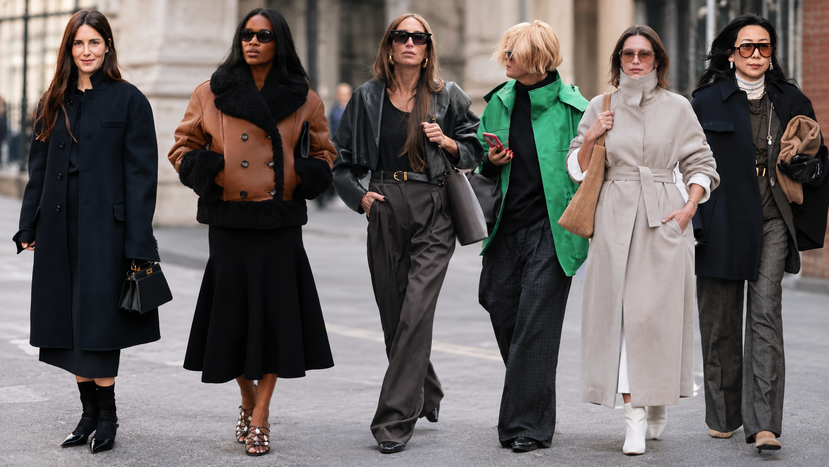 Four women in layered fall outfits, including sweaters, skirts, jeans, scarves, and loafers, walk along a city street.