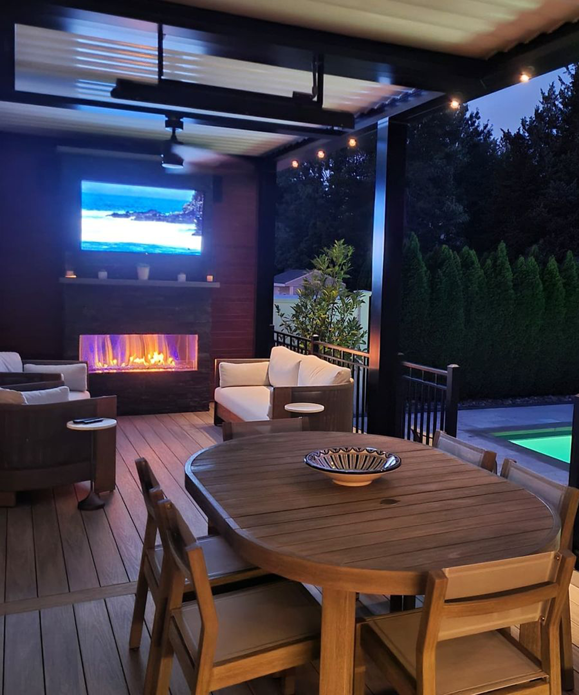 Deck idea pictured in evening with electric fireplace, dining table, sofas and TV