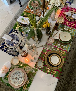 A birds eye view of a dining table with a white tablecloth and coloful placemats with vintage plates, gold cutlery, glass candlesticks, plants, lemons, and white flowers on it