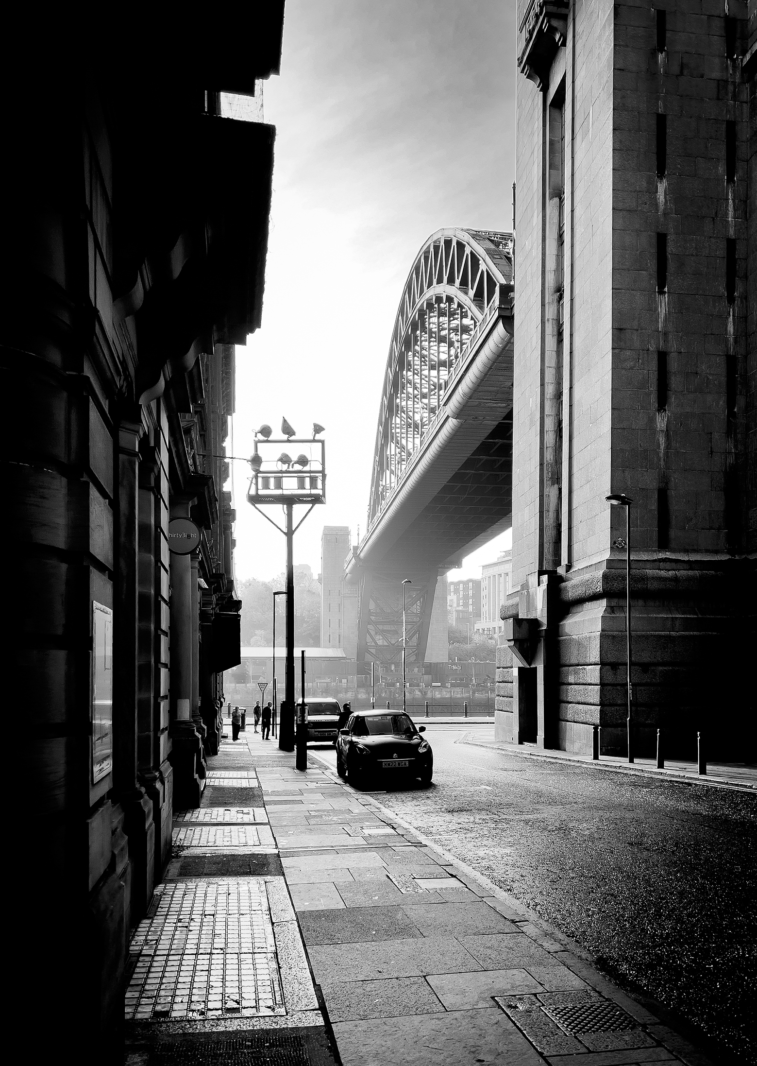 A black-and-white street scene showcasing the Tyne Bridge above, flanked by historic buildings and scattered pedestrians
