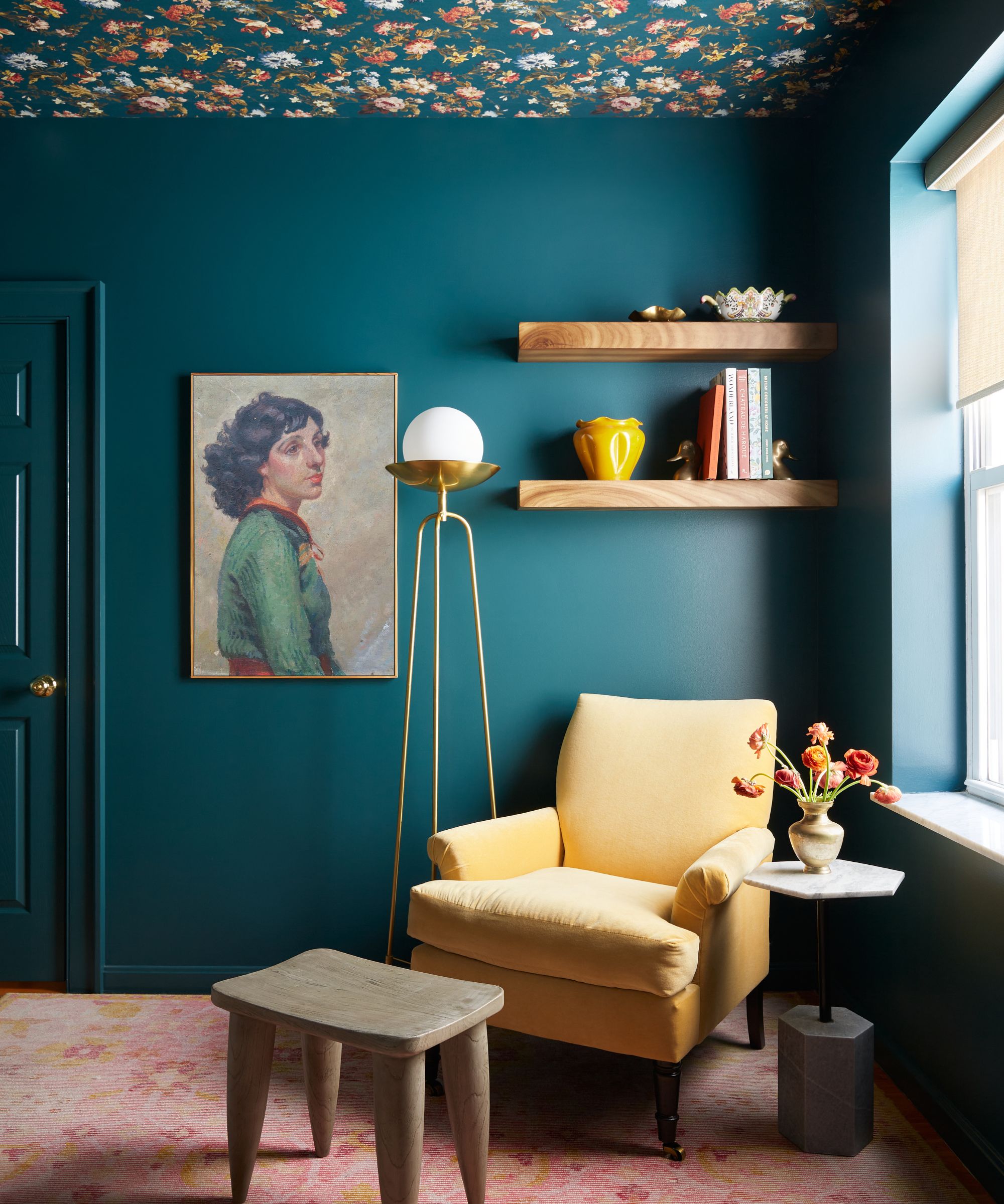 A home office with dark teal walls, a floral wallpapered ceiling, a pink carpet, a mustard armchair, and portrait artwork on the wall.