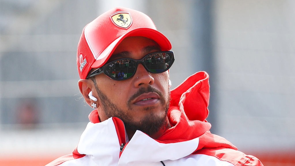 Lewis Hamilton, wearing Ferrari tracksuit and cap, looks on ahead of a Grand Prix