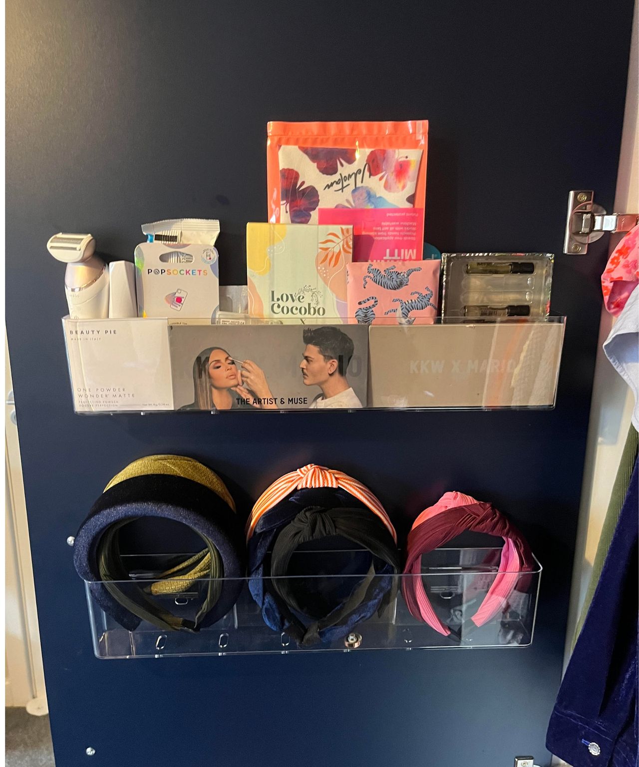 Front on view of my two clear plastic adhesive organizers in my matte dark blue closet door. The top one is filed with narrow makeup and cosmetic pads and the lower one holds 6 headbands in various colors