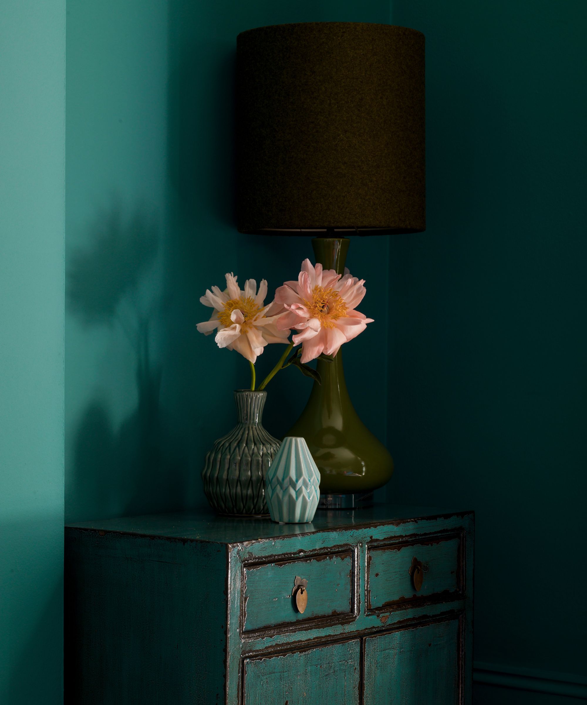 A green cabinet with two green vases with pink flowers and a green vase with a black lampshade. Behind are green walls.