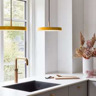 white kitchen corner with basin, white worktops, vase of dried flowers, brass tap, pair of matching yellow modern pendants above the sink