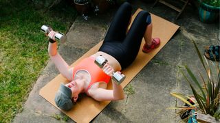 Woman lying on back on yoga mat holding dumbbells doing a chest press