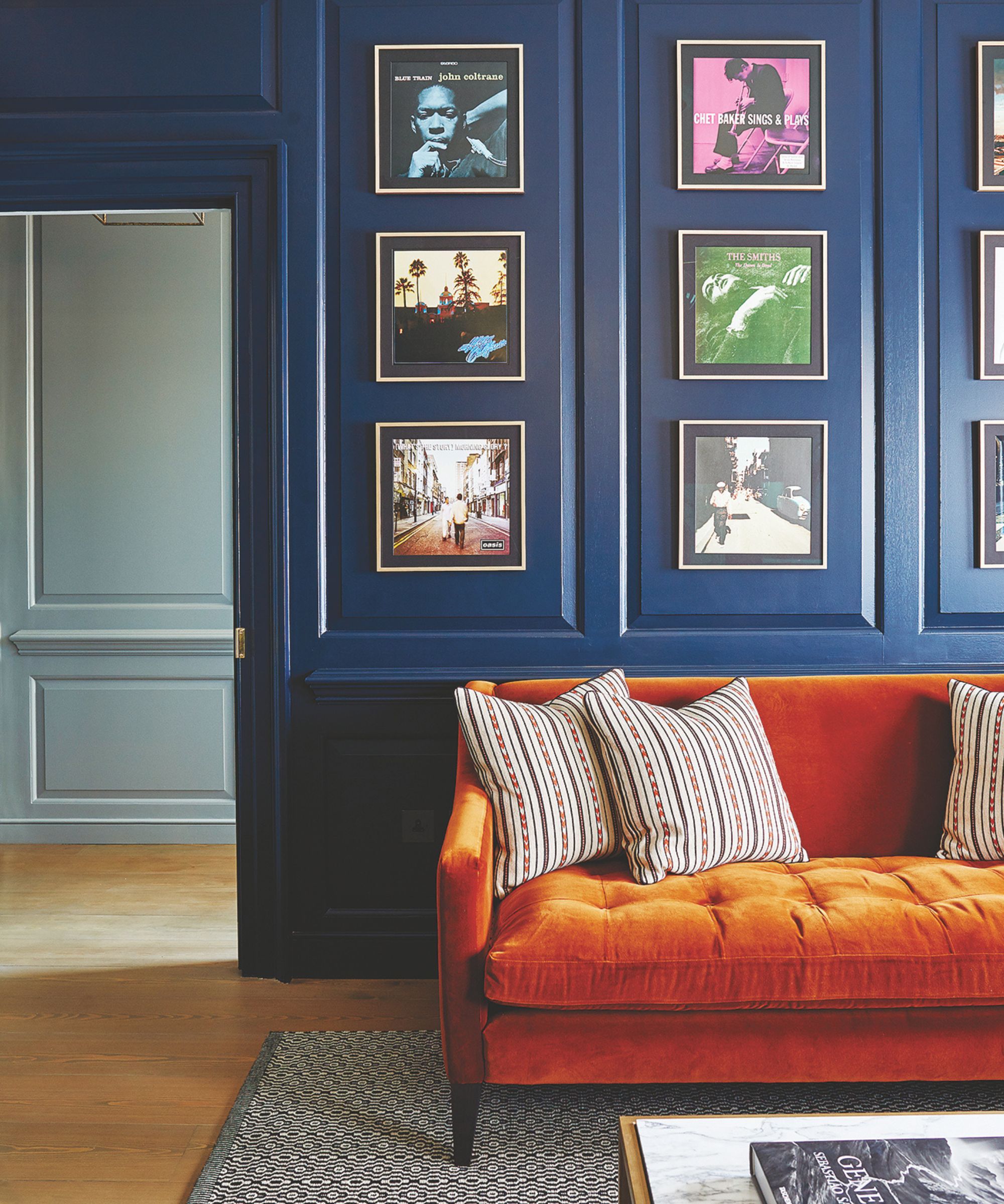 A dark blue panelled living room with a burnt orange sofa with striped cushions seen to the right. Beneath the couch is a black and white patterned rug, and in front of it is a white marble and gold coffee table with a coffee table book on top. Above the sofa, on the wall, are several framed records, and to the left a room beyond is seen with light blue panelled walls. To flooring in both is light brown wood.