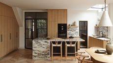 A mid tone wood kitchen with richly veined marble countertop and island, wishbone bar chairs and a white cone-shaped pendant over the dining table
