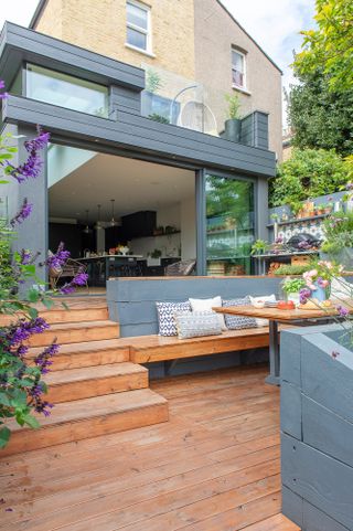 Modern garden ideas: a decked garden on two levels with grey painted raised planters and natural stained wooden flooring, leading into an open-plan kitchen