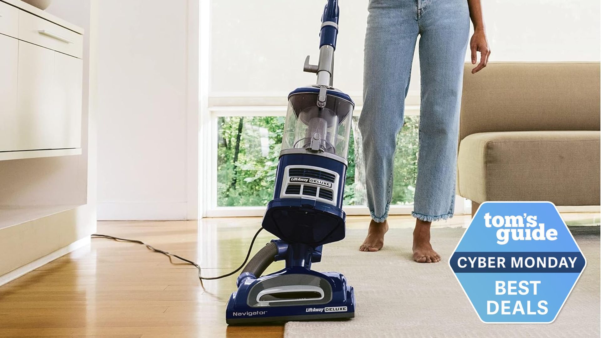 A woman's legs in a beige living room as she pushes a Shark Navigator vacuum cleaner across a carpet and wooden floor