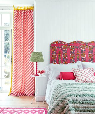White bedroom with pink patterned headboard, white bedding with green throw, pink scatter cushions, white side table with red and green table lamp, floor to ceiling window with pink striped curtains, looking out onto garden, pink patterned rug
