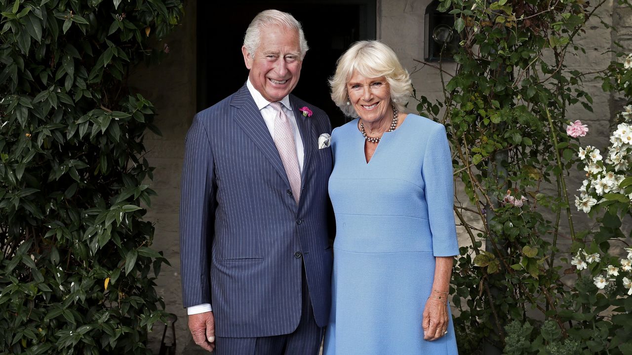 King Charles wearing a blue suit standing next to Queen Camilla, wearing a blue dress, in a garden outside 