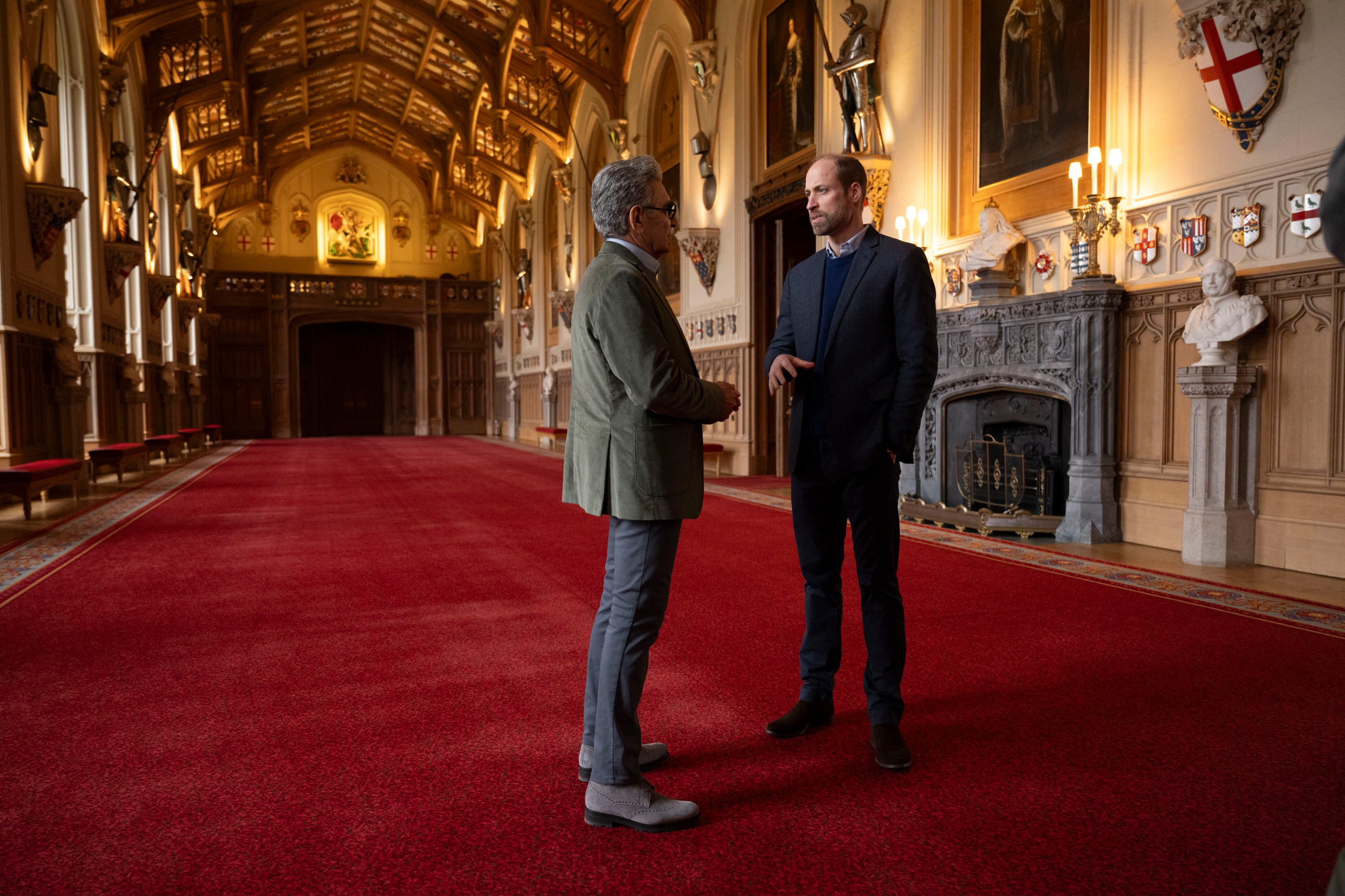 Prince William and Eugene Levy talking in a red carpeted hall at Windsor Castle