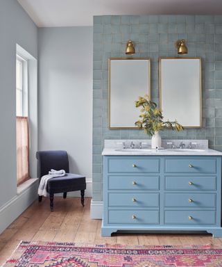 Blue painted bathroom with blue tiled feature wall, blue wash stand with two sinks, two mirrors and two wall lamps, pink rug and chair in corner of the room