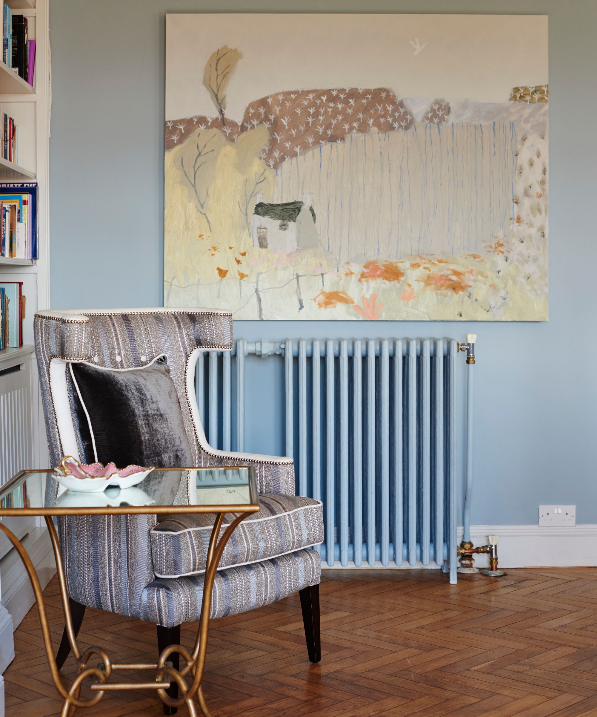A blue room with white bookshelves, a striped grey and purple striped chair, gold and glass table, a large landscape artwork and an umbrella painted the same color as the walls.