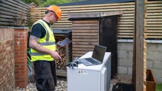 HVAC worker in protective clothing checking an air source heat pump on his tablet and laptop, the pump is installed outside in the garden.