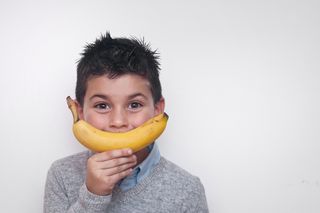 A young brunette boy holding a banana over his mouth to symbolise a smile