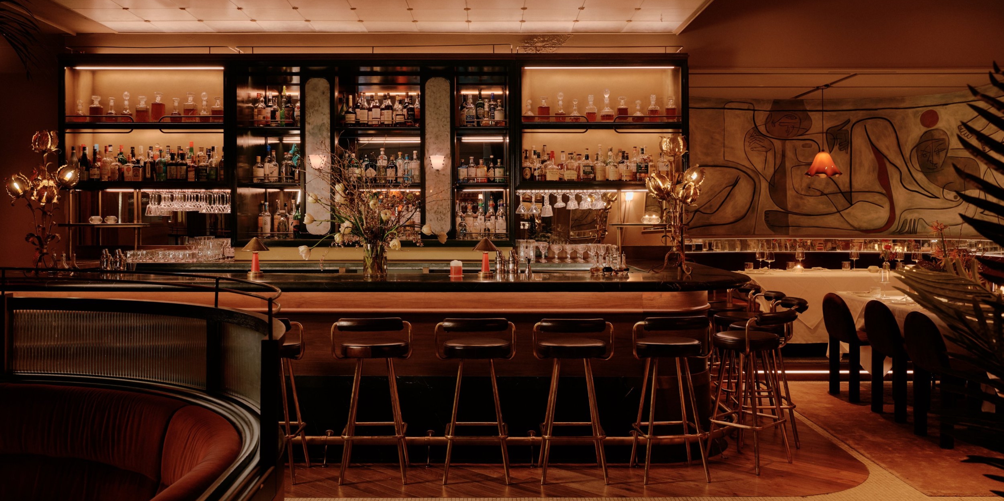 A contemporary take on Art Deco-style interiors brings a warmly lit, dazzling cocktail bar to life through plush wood, velvet, and leathery surfaces, thriving plants, and sculptural lighting.