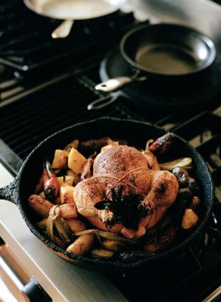 Close up of a black frying pan with a whole chicken and vegetables being cooked on a hob, empty pans to the back of the shot