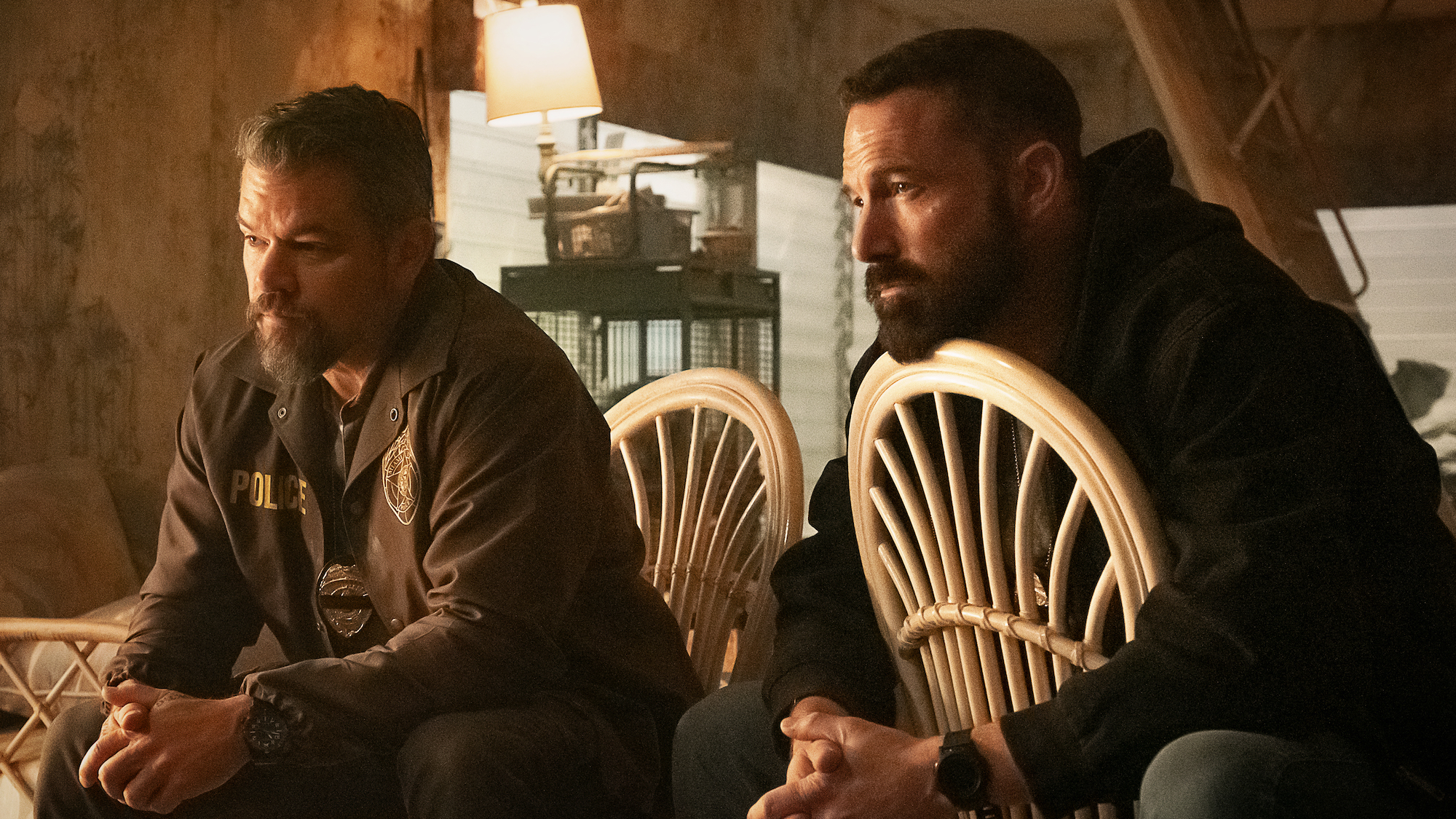 Ben Affleck and Matt Damon sitting next to each other in wooden chairs and in cop uniforms in The Rip
