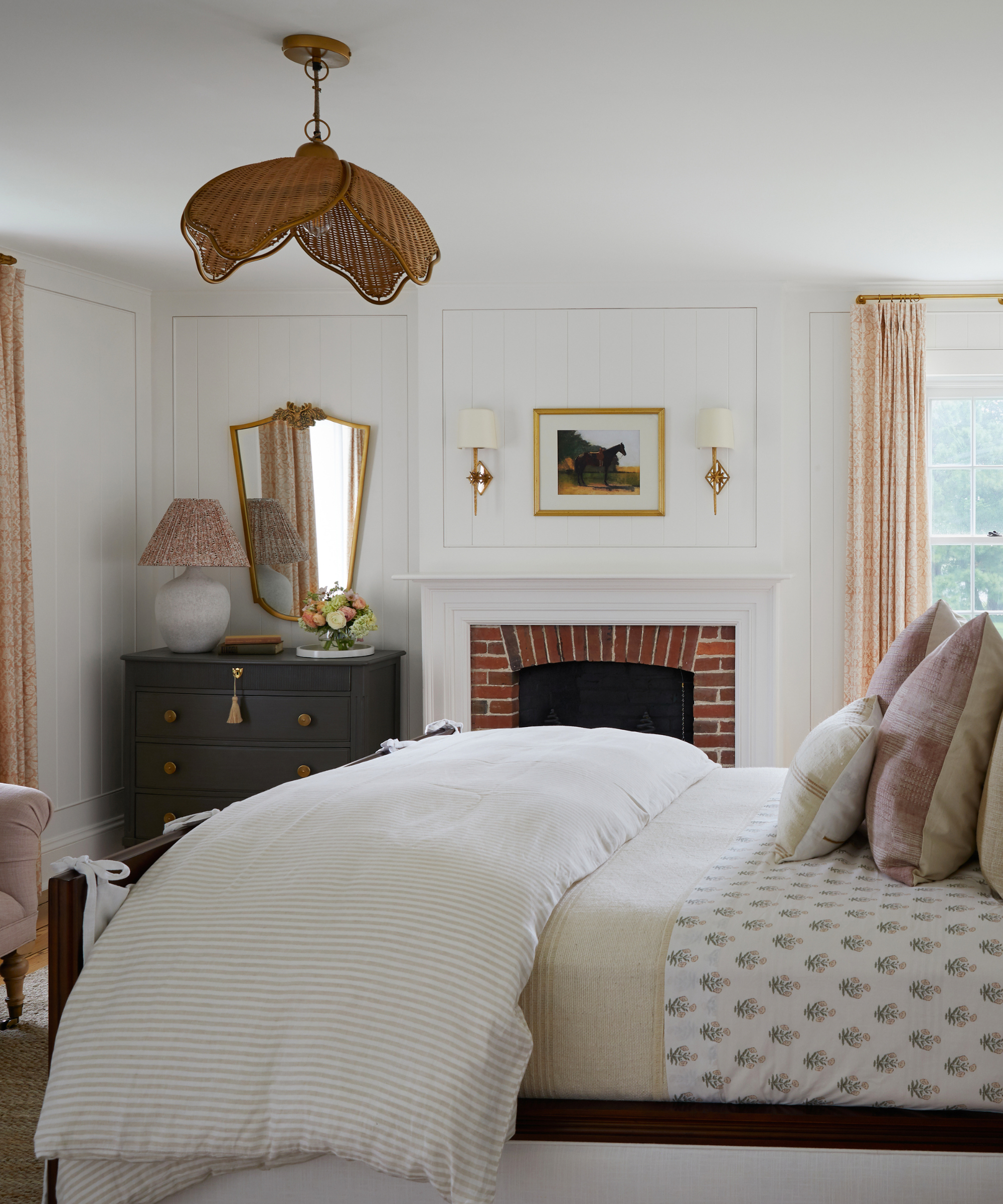 a bedroom with white walls and a white ceiling, a large bed with a down comforter and many pillows, a black dresser in the corner next to a fireplace, and a rattan ceiling light