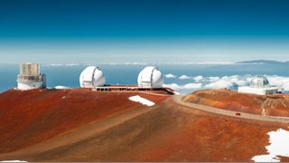 Keck Observatories sit atop rusty red terrain and a blue sky above.
