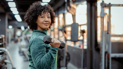 A woman stands smiling in a gym and a holding a dumbbell in her right hand.