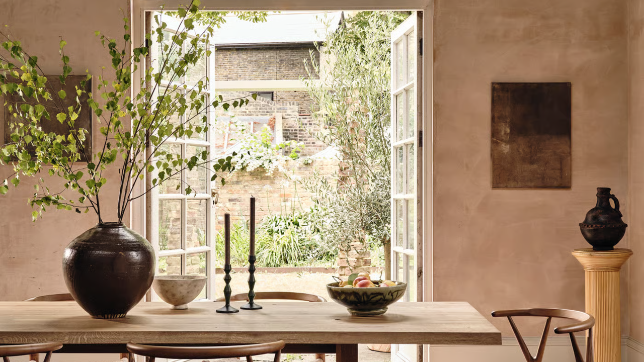 A dining room with limewashed walls and a dining table with a large glass of foliage on top, two taper candles and two bowls, with open patio doors leading out to a courtyard 