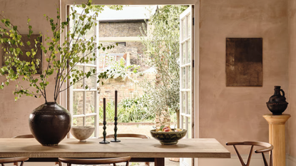 A dining room with limewashed walls and a dining table with a large glass of foliage on top, two taper candles and two bowls, with open patio doors leading out to a courtyard 