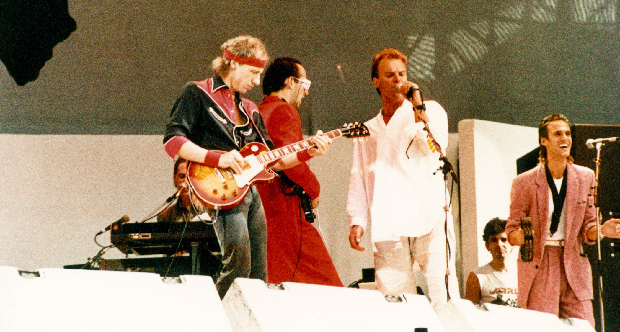 Dire Straits perform at Live Aid 1985, with Sting joining the band onstage at Wembley for Money for Nothing. Sting wears a white suit. Knopfler has a red-and-black western shirt, a red headband, and a Gibson Les Paul Standard.