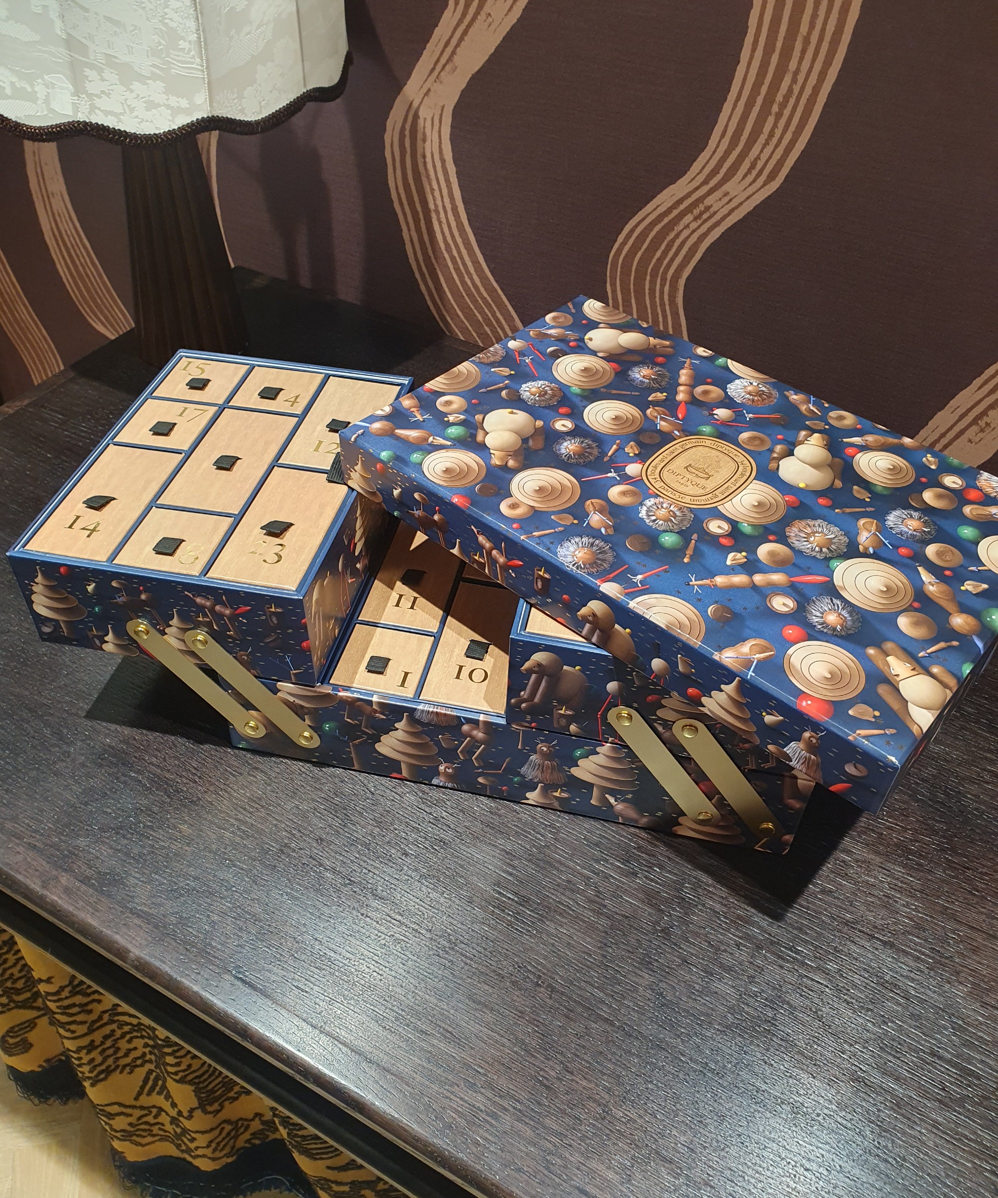 An open Diptyque luxury advent calendar for 2026 set on a dark wood table with a brown patterned wallpaper in the background