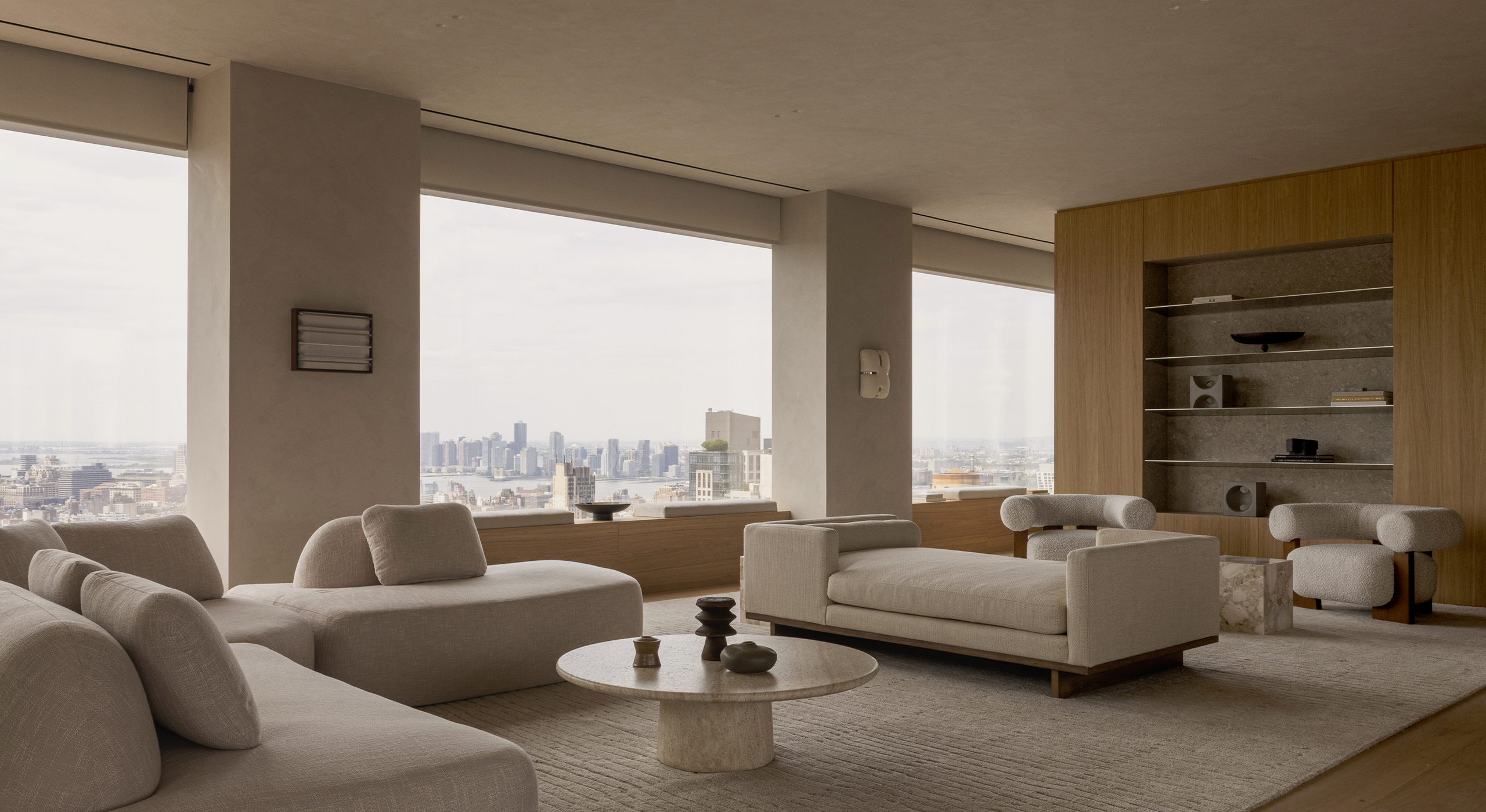 Neutral-toned living room with floor-to-ceiling windows overlooking the Manhattan skyline, featuring sculptural seating, natural oak cabinetry and a stone coffee table.