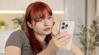 Young woman at home scrolls news on smartphone feels bored and dissatisfied and low energy. Redhead girl on sofa reads feed long time tired eyes slow breath shows emotional exhaustion and deep sigh.