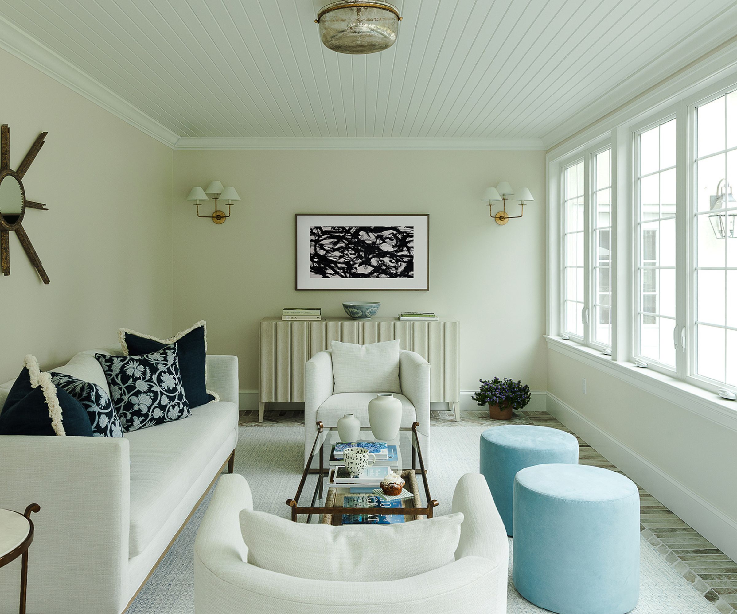 All-white sunroom with large windows along the right wall, white shiplap ceiling, cream sofa with navy blue floral cushions, two pale blue ottomans, cream armchairs, and cream rug.