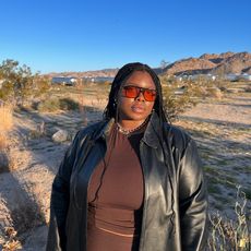 Chichi wears brown skirt and tank set with leather jacket and aviator sunglasses in the desert