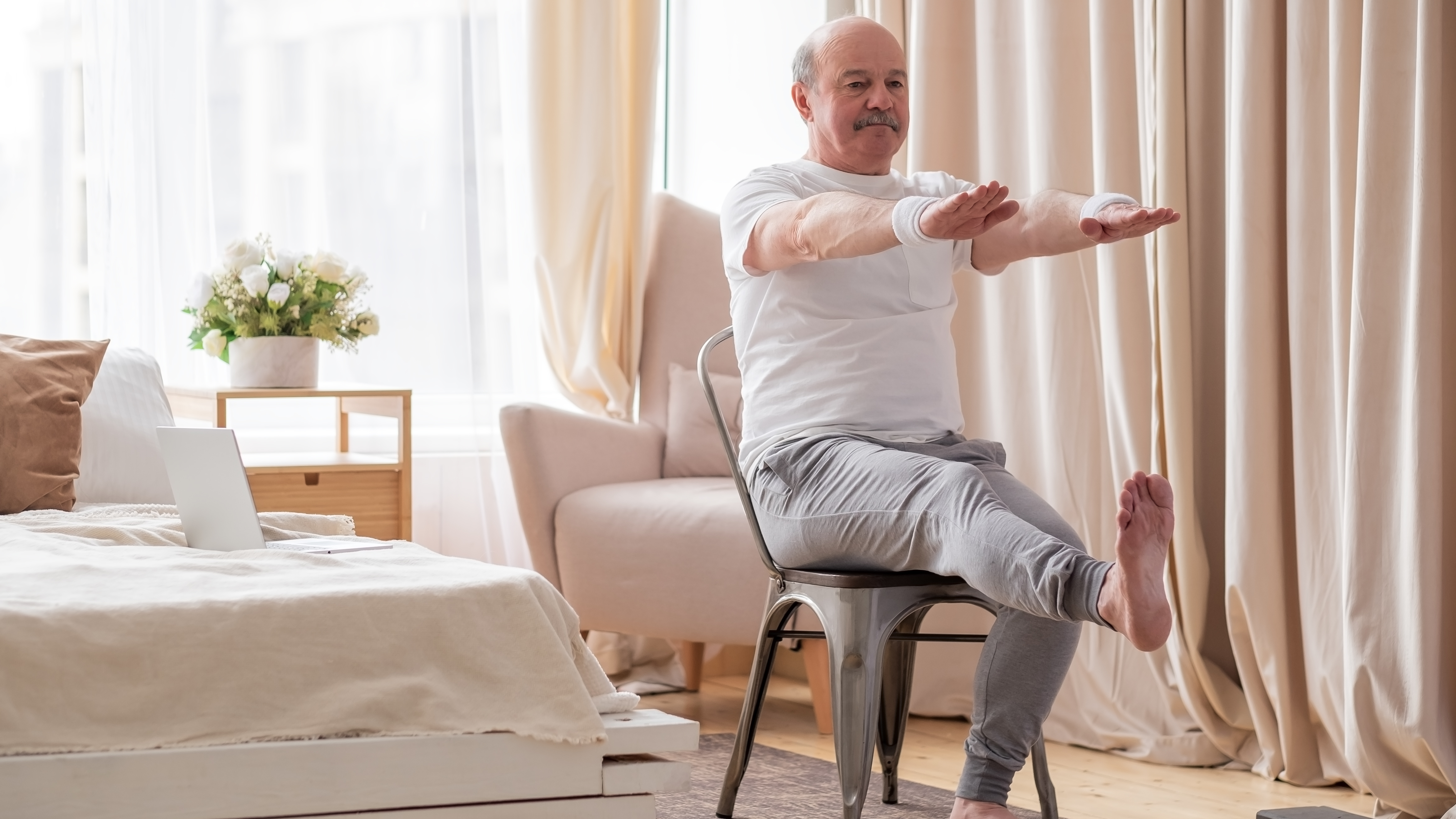 A man exercises in his bedroom on a chair. He is sitting with his arms straight in front and his right leg extended, so it's parallel to the floor. We see a bed, nightstand and armchair behind him.