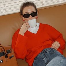 Model wearing white t-shirt, jeans, and red Almina Concept v-neck sweater, sitting on couch and drinking out of coffee cup.