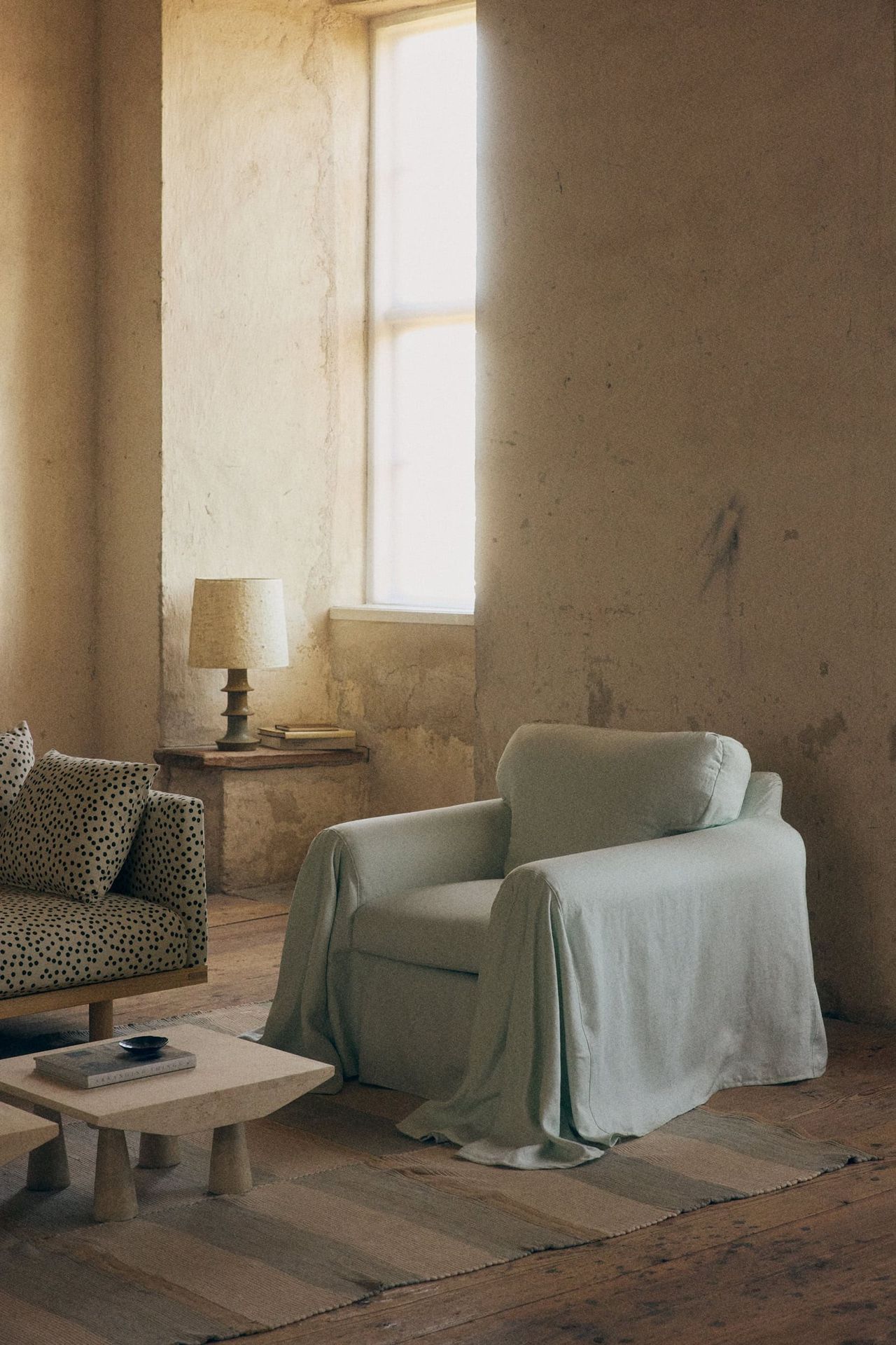 Image of a vintage living room with unfinished-looking plaster walls, a light blue accent chair with a draping cover on it, a blue and white striped rug, and a polka dot sofa in view.
