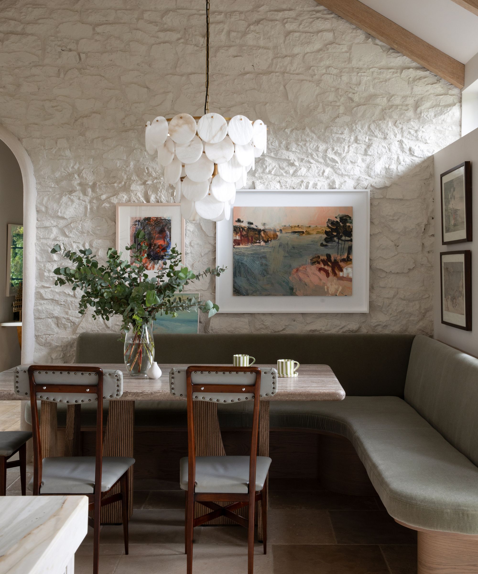 A kitchen seating area with an L-shaped green banquette, a wooden table with a marble top, and exposed stone walls washed in white and covered in vintage art