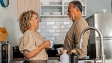 An older couple smile and talk as they face each other in their kitchen.