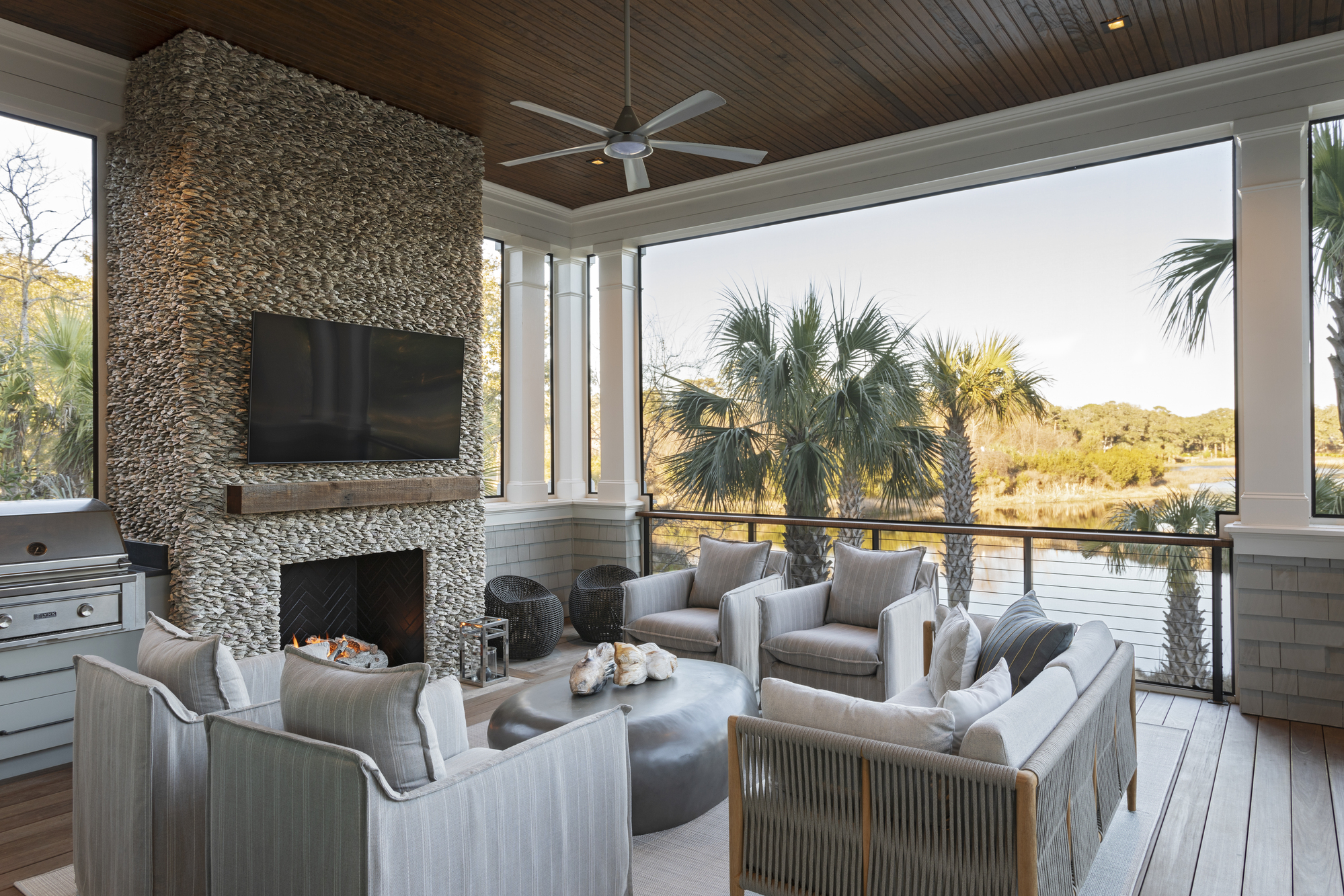 outdoor living room with oyster fireplace with TV, fan on timber ceiling, deck with four armchairs and one rope-style outdoor sofa, round coffee table on rug, and views to marshland