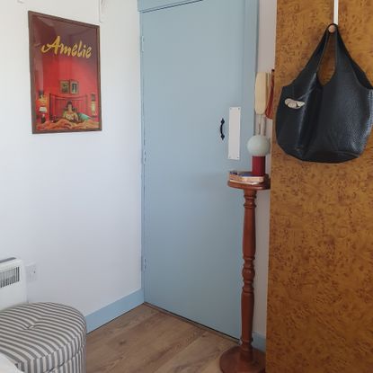 Sara's entrance area with a striped storage ottoman, a side table with a lamp next to the front door and a framed film poster hanging on the wall